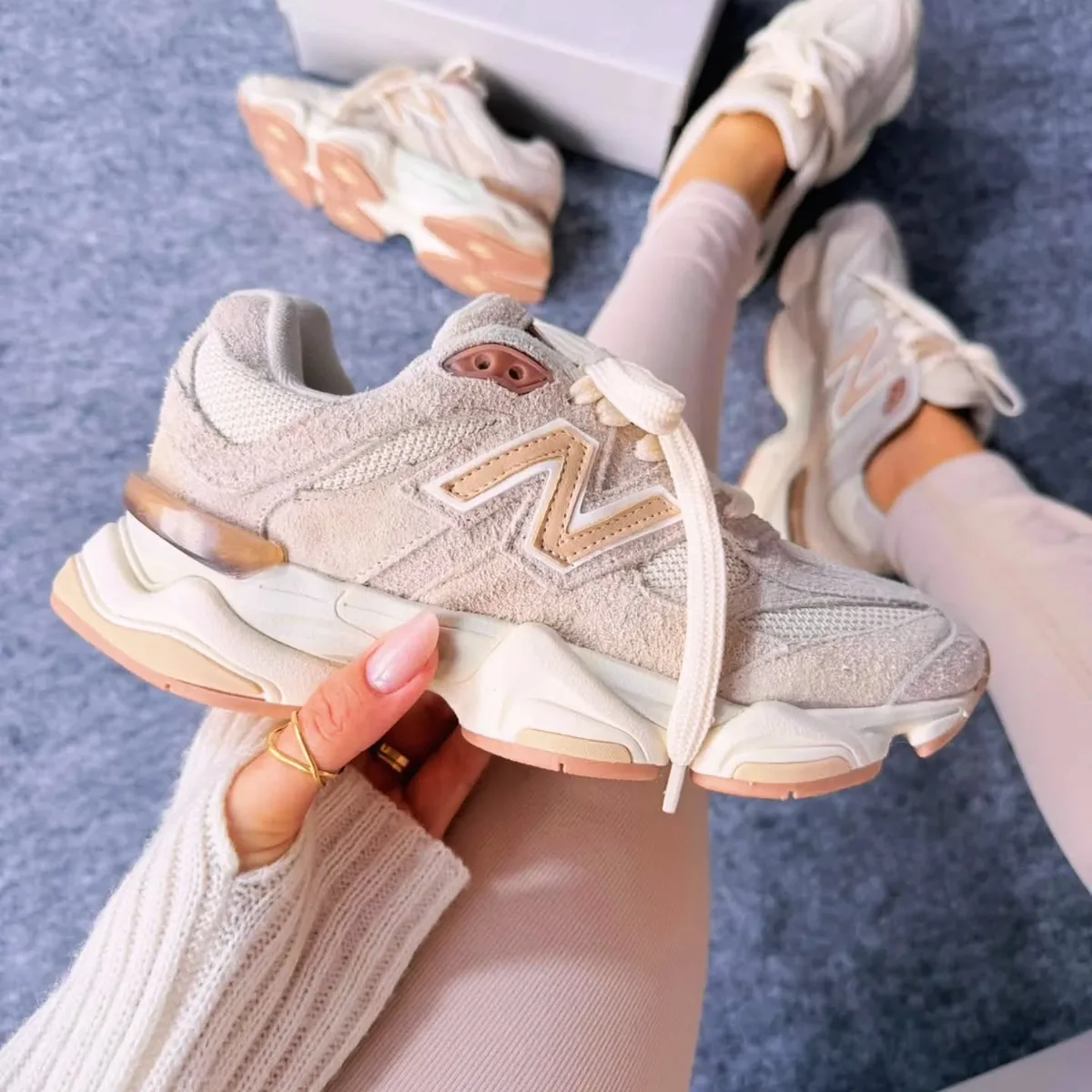 Uma mão de unhas feitas segurando o New Balance 9060 areia e rosé, sobre o tapete cinza