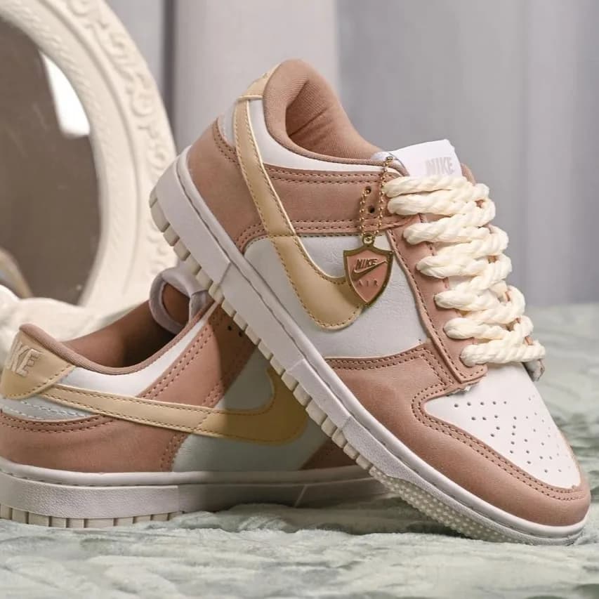 O Nike Dunk Low rosé e dourado, com cadarço creme e pingente, sobre a manta branca